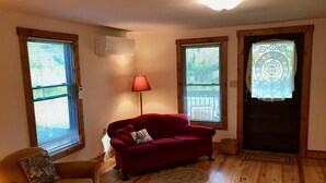 A living room with a red sofa, a yellow armchair, and a lamp. There are three windows with blinds, a wooden floor, and a welcome mat.