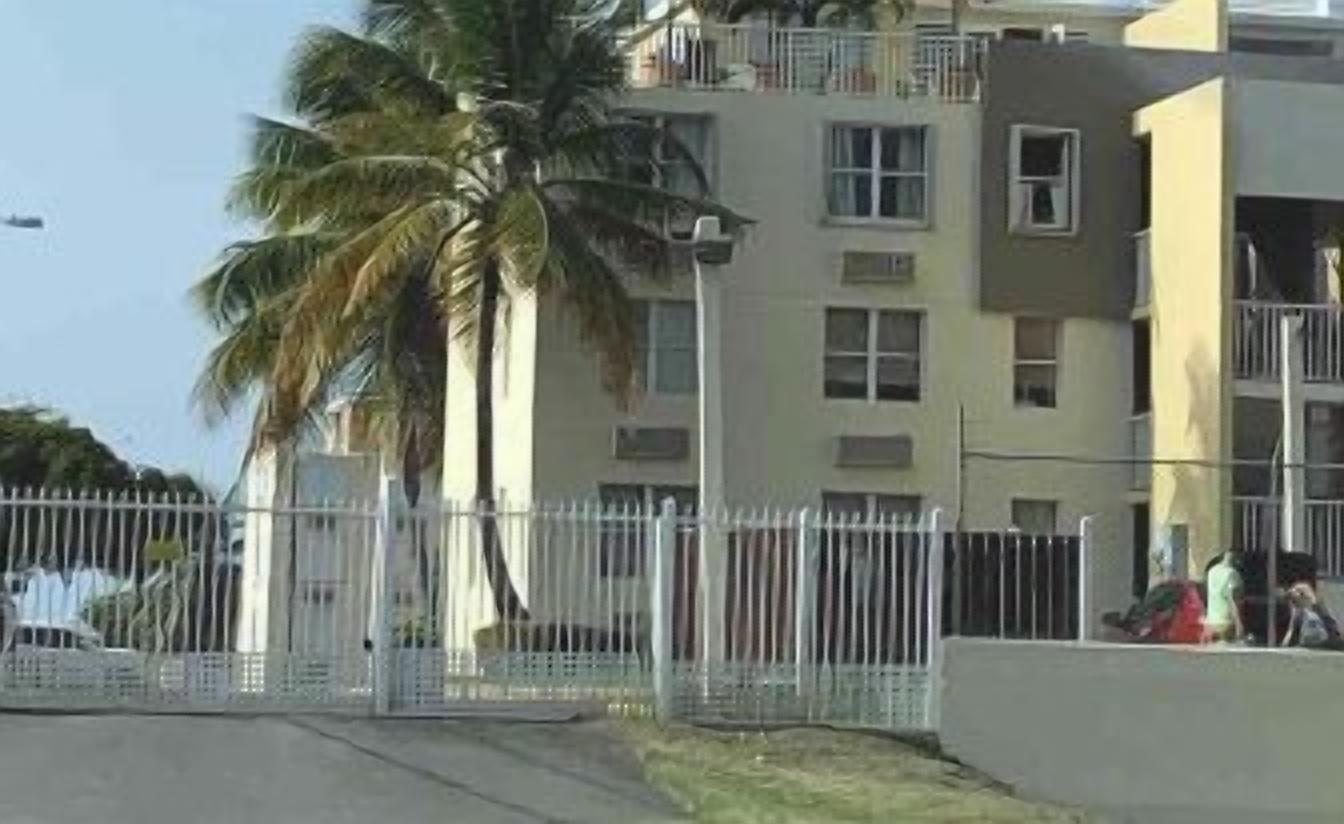 Un edificio residenziale a più piani con balconi, un recinto bianco e una palma sullo sfondo.