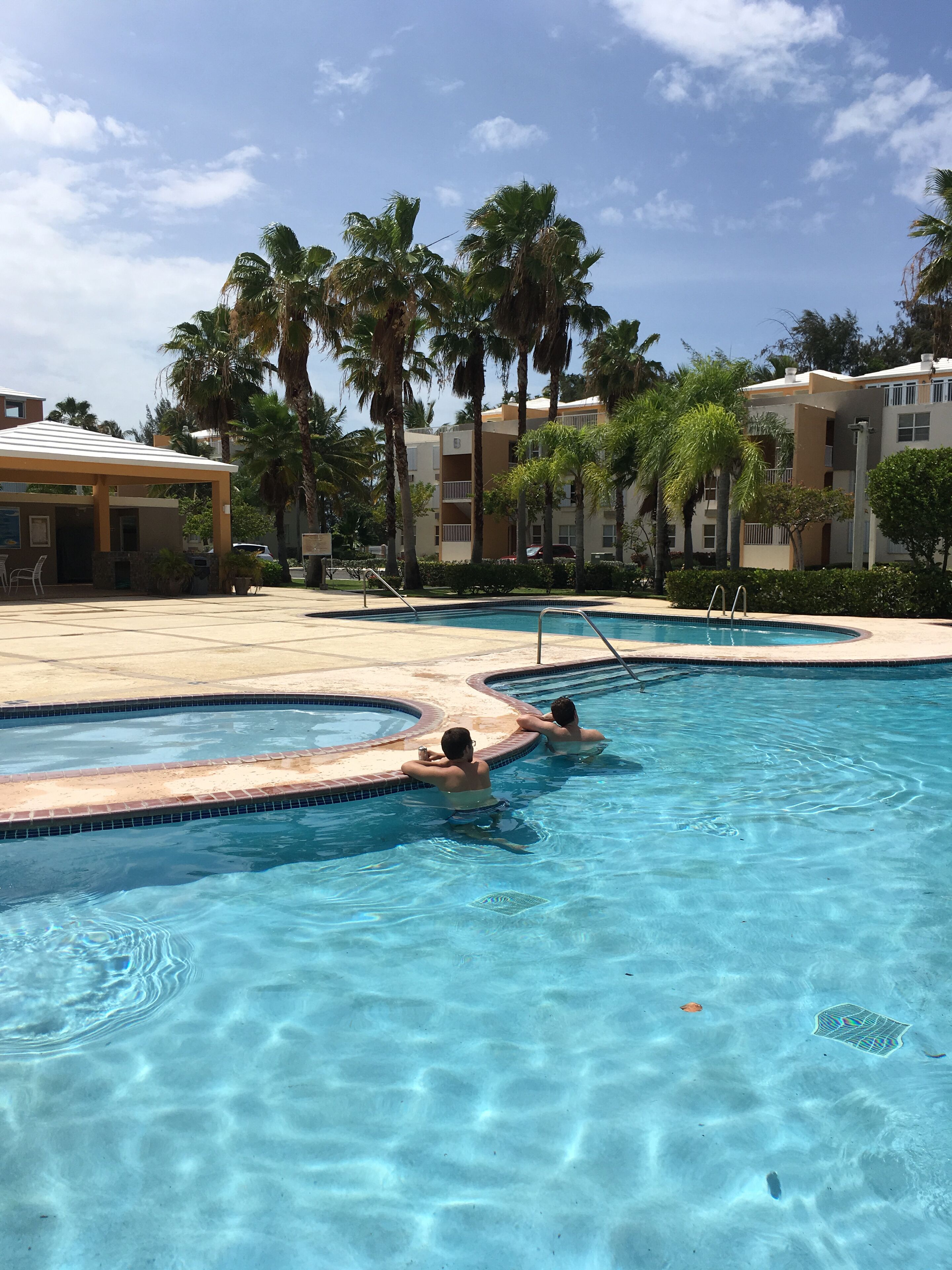 A clear blue pool with two people swimming, surrounded by palm trees and a building with balconies.