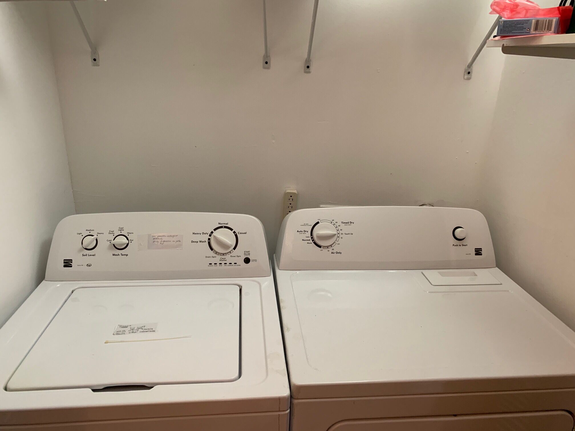 Deux machines à laver à chargement frontal se trouvent dans une buanderie, avec un mur blanc et une étagère au-dessus.