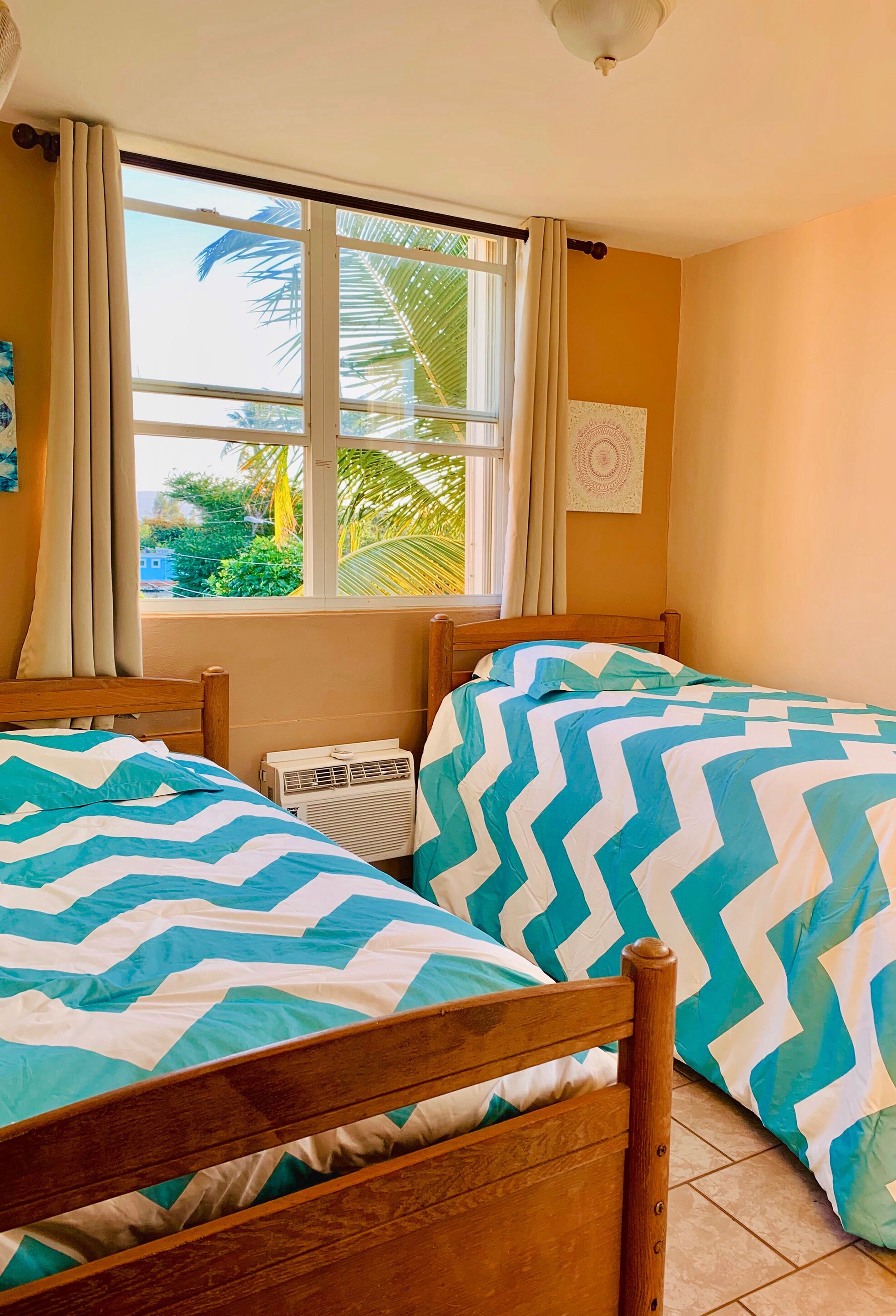 A room with two wooden beds, a window with curtains, an air conditioning unit, and a view of greenery outside.