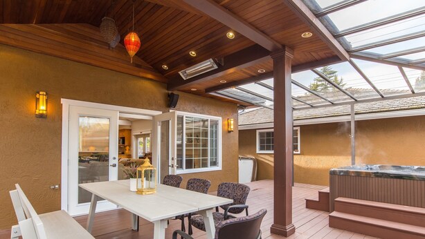 An outdoor patio with a wooden table and chairs, a hot tub, and a covered area with a glass ceiling.