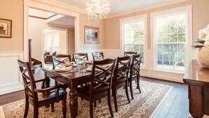 A dining room with a large wooden table, six chairs, a chandelier, and large windows with a view of greenery.