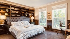 A bedroom with a large bed, wooden shelving, a small table, and a window with a view of greenery.
