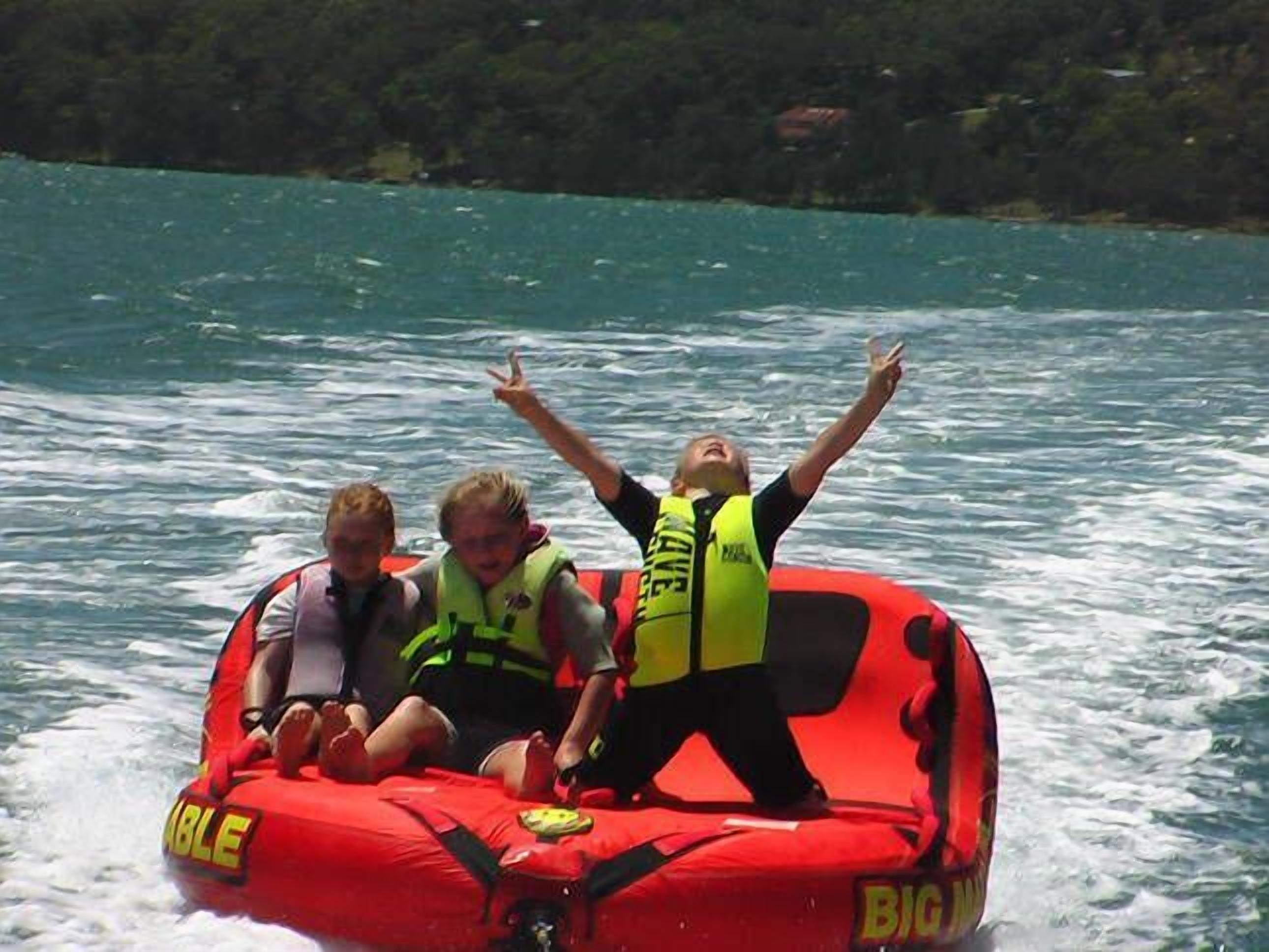 A red inflatable boat with two people wearing life jackets and one person standing with arms raised.