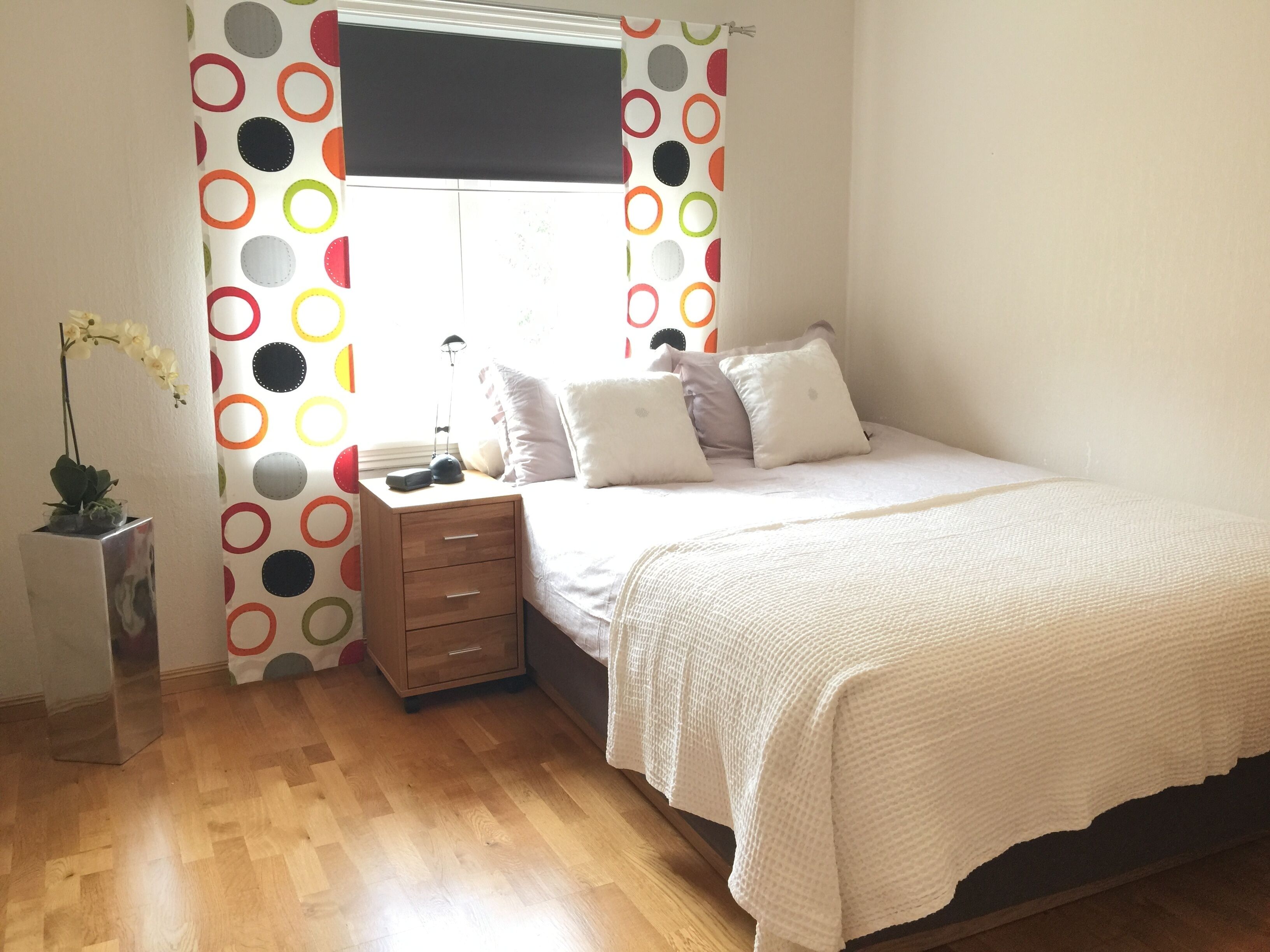 Quarto com cama, mesa de cabeceira, abajur e janela com cortina de bolinhas.