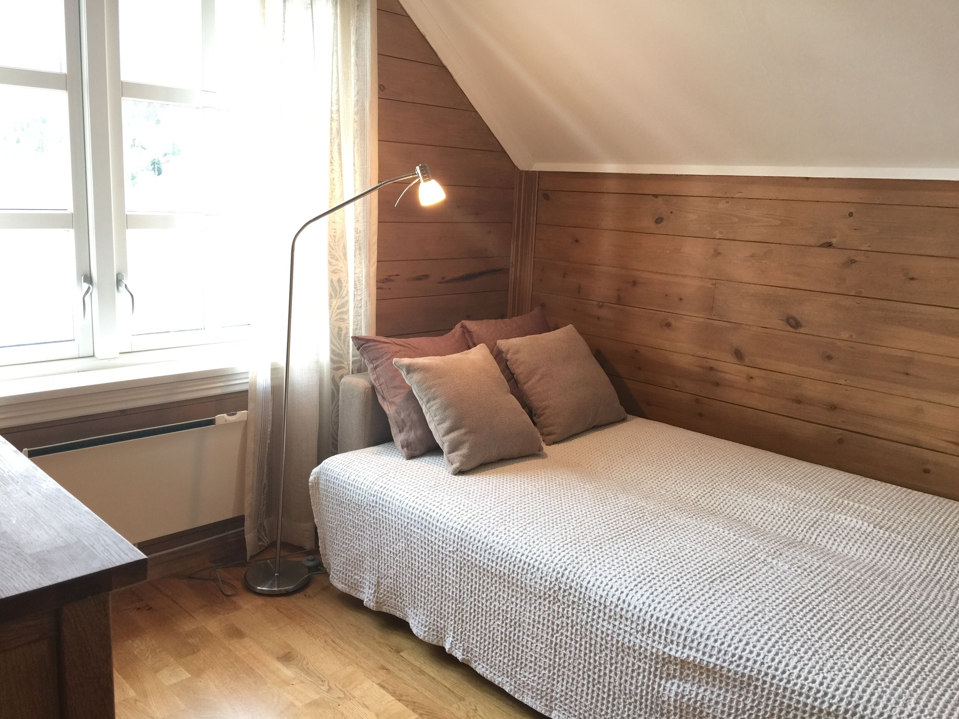 Una habitación acogedora con techo de madera, una cama con manta tejida, una lámpara de pie y una ventana con cortinas.