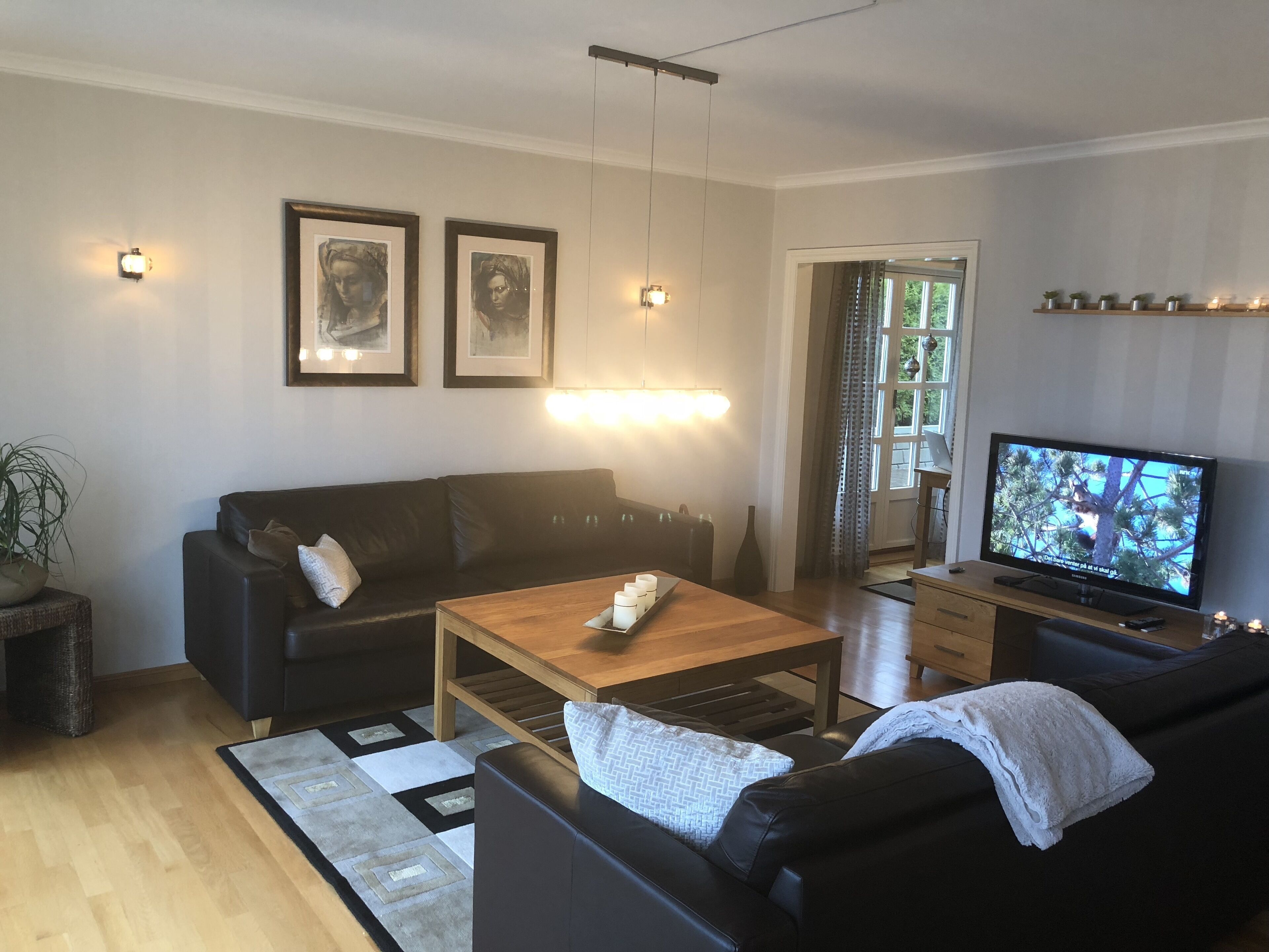 Ein Wohnzimmer mit einem schwarzen Ledersofa, einem Holztisch, einem Fernseher auf einem Schrank und gerahmten Bildern an der Wand.