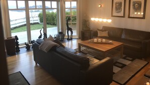 A modern living room with a large window, a black leather sofa, a wooden coffee table, and a view of a deck and greenery outside. - Charming House with a Fantastic View of Norway's Nature (Ørsta)