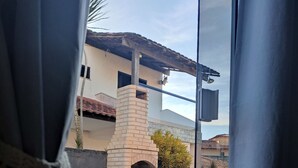 Blick durch ein Fenster mit Vorhängen, der einen Ziegelgrill im Freien, weiße Kunststoffstühle und einen Tisch zeigt.