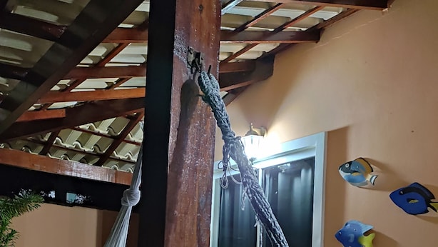 Eine Hängematte, befestigt an einem Holzbalken in einem Raum mit Fliesenboden, einem Fenster und Wandbildern mit Fischmotiven.