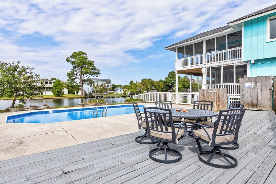 A poolside area with a wooden deck, a table with chairs, and a building with a balcony.