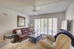 A living room with a leather sofa, a glass coffee table, a beige armchair, a ceiling fan, and a large window with blinds. - Beach Club 207, Oceanfront complex (St Simons Island)
