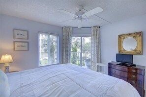 A bedroom with a bed, a ceiling fan, a television on a dresser, a window with curtains, and framed pictures on the wall. - Beach Club 207, Oceanfront complex (St Simons Island)