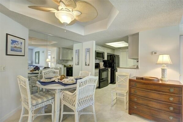 A dining area with a table set for four, white wicker chairs, a ceiling fan, and a wooden dresser with a lamp.