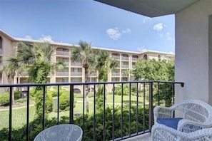 A balcony with white wicker chairs, a view of a building with multiple balconies and palm trees, and a well-maintained lawn.