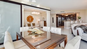 A modern living room with a glass coffee table, white sofas, and a dining area with a chandelier.