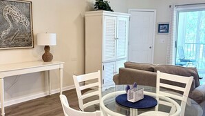 Un coin repas avec une table en verre et des chaises blanches, un meuble blanc, un ventilateur de plafond et une vue à travers une porte vitrée.