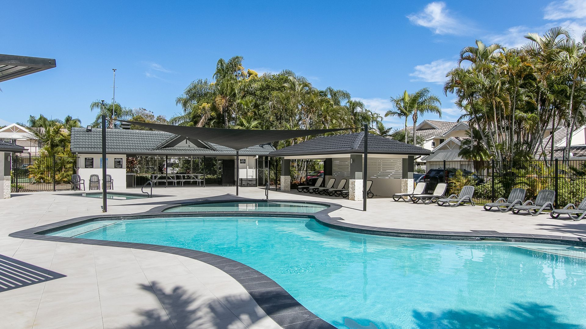A swimming pool with lounge chairs and a covered seating area, surrounded by palm trees and houses.
