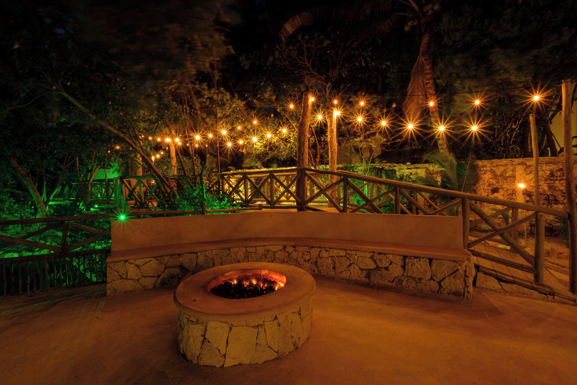 A stone fire pit surrounded by a wooden deck with railings, set against a backdrop of trees and string lights.