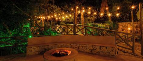 A stone fire pit surrounded by a wooden deck with railings, set against a backdrop of trees and string lights.