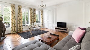 A spacious living room with a large sofa, a coffee table, and a TV. There are large windows with curtains, a chandelier, and a statue. - CAVALIER DES LIONS 2bedr 2 1/2baths (Paris)