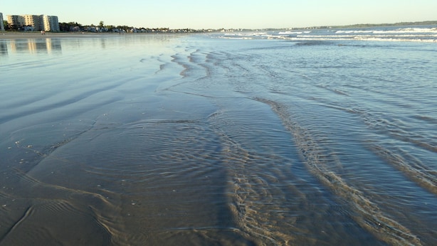 A beach with clear water and gentle waves, buildings in the background, and a clear sky.