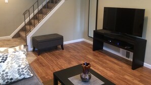 Una sala de estar con piso de madera, una escalera, un mueble negro para entretenimiento, un televisor de pantalla plana y un florero sobre una mesa de centro.