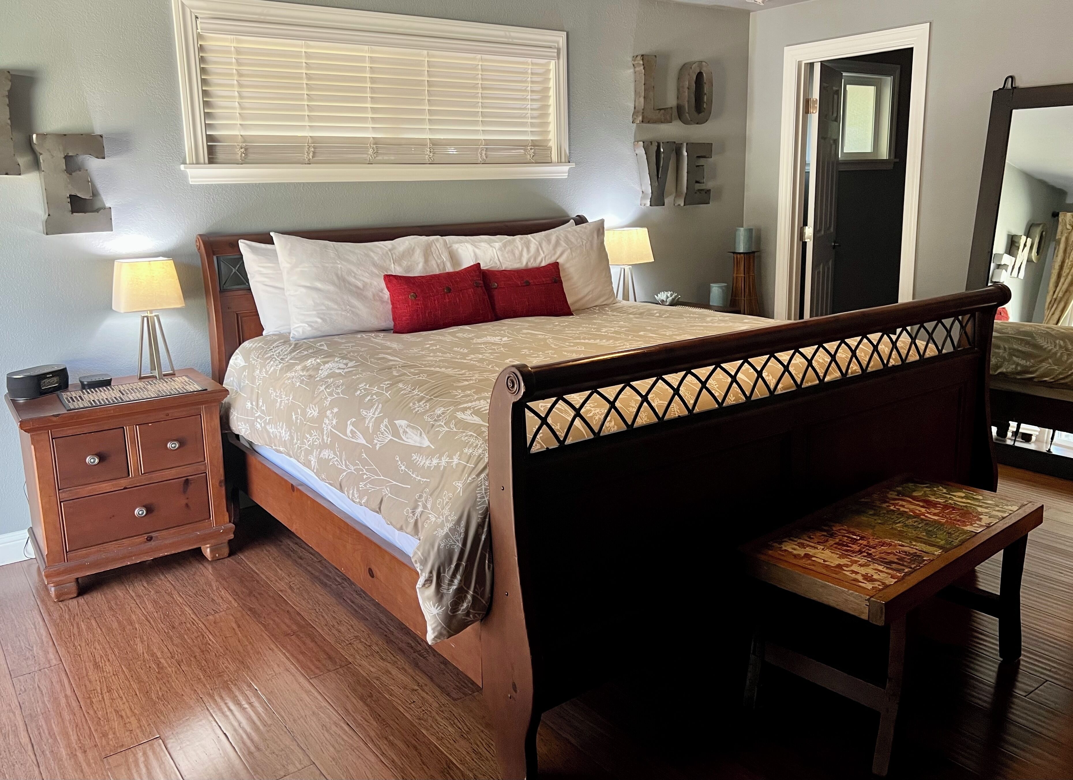 Un dormitorio con una cama de madera, dos mesitas de noche, un banco, una ventana con persianas y un espejo.