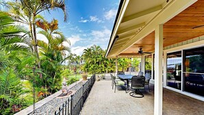 Un patio techado con suelo de ladrillo, mobiliario exterior y vistas a plantas tropicales y un cielo despejado.