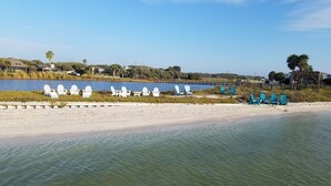 A lakeside view with chairs and tables on the shore, a sandy beach, and houses in the background.