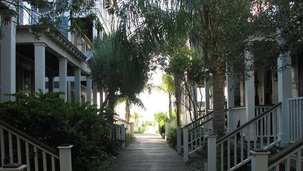 A wooden pathway flanked by white railings and lush greenery, leading towards a building with multiple windows and balconies.