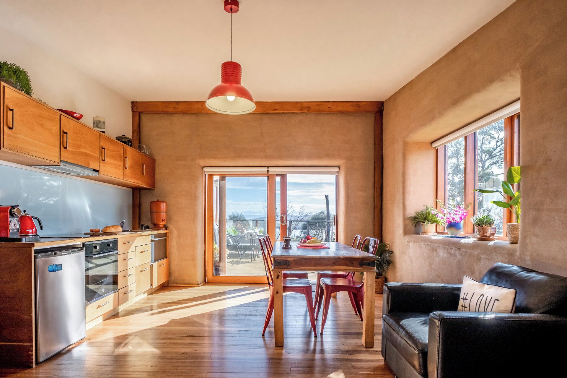 Une cuisine spacieuse avec des meubles en bois, une table à manger avec des chaises, un canapé en cuir noir et une grande fenêtre agrémentée de plantes.