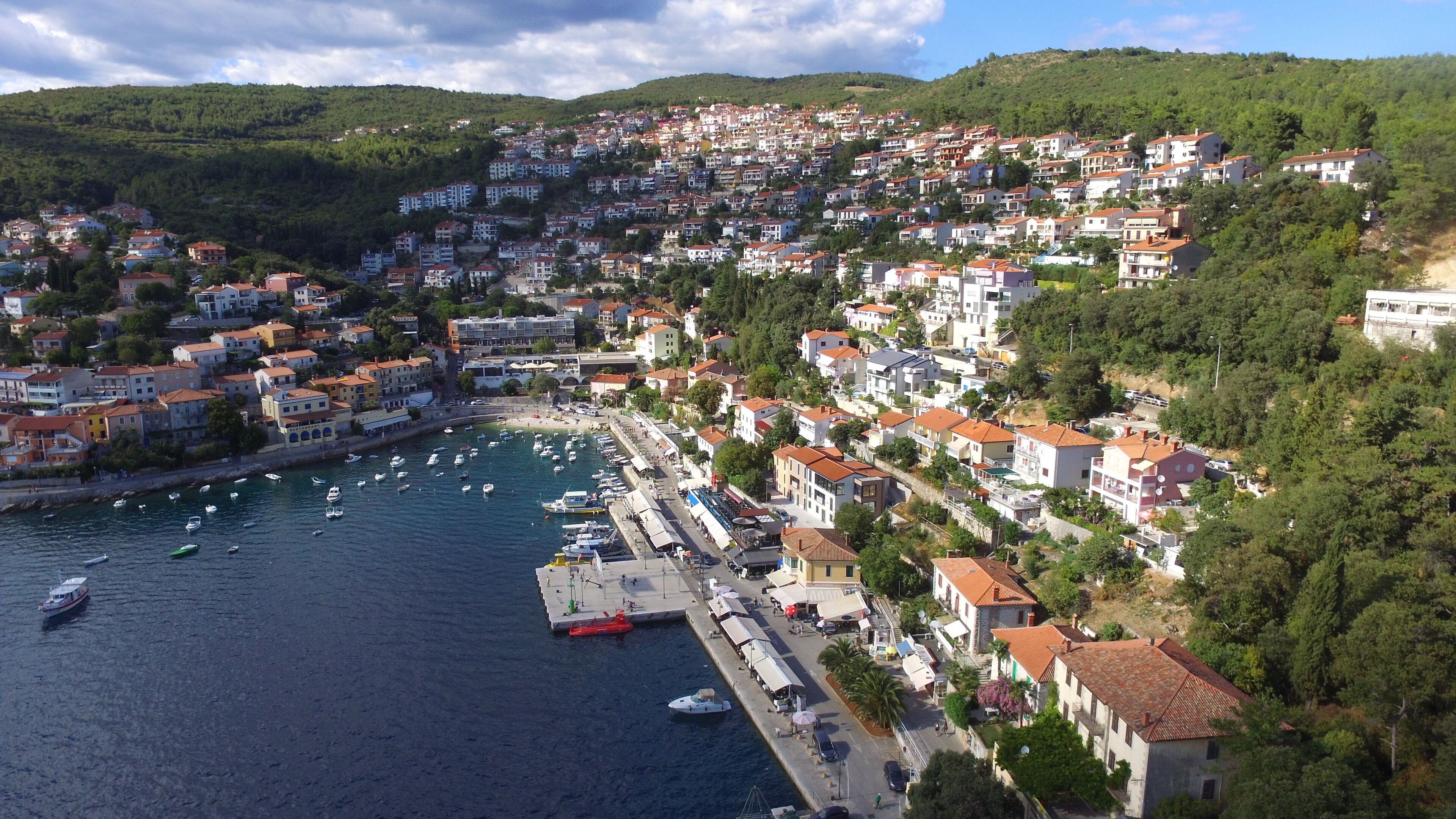 A coastal town with buildings on a hillside, a marina with boats, and a waterfront promenade.