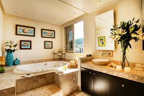 A modern bathroom with a large bathtub, a sink, and a vanity. There are framed pictures on the wall, a vase with flowers, and a window providing a view of the outside. - Panoramic Oceanview, Luxury Condo 3 bd, 3bath on the beach. (Puerto Vallarta)