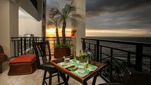 A balcony with a table set for two, a wicker chair, a potted plant, and a view of the ocean at sunset. - Panoramic Oceanview, Luxury Condo 3 bd, 3bath on the beach. (Puerto Vallarta)