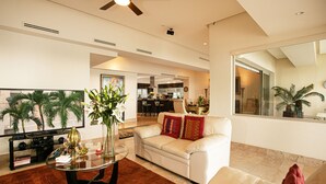A modern living room with a white sofa, a glass coffee table, and a TV. There is a kitchen in the background with a dining area. - Panoramic Oceanview, Luxury Condo 3 bd, 3bath on the beach. (Puerto Vallarta)