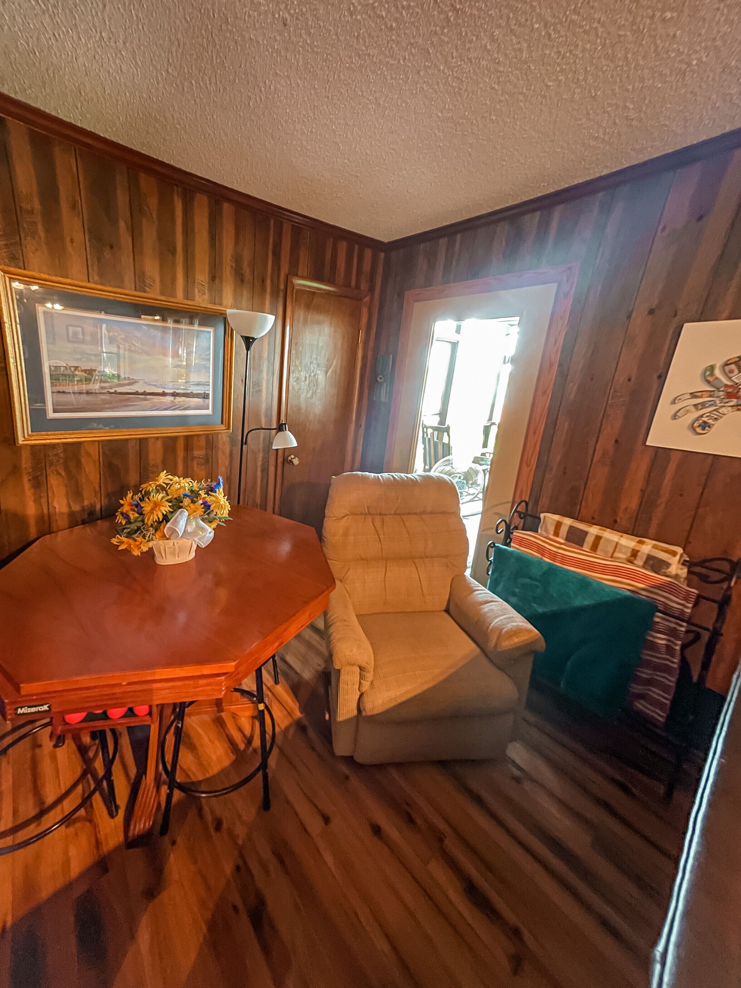 Una habitación con paneles de madera, una mesa de comedor, una silla acolchada, una lámpara de pie y un cuadro en la pared.