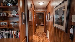 A wooden-paneled hallway with bookshelves, framed pictures, and a door leading to another room. - Chechessee River House (Okatie)