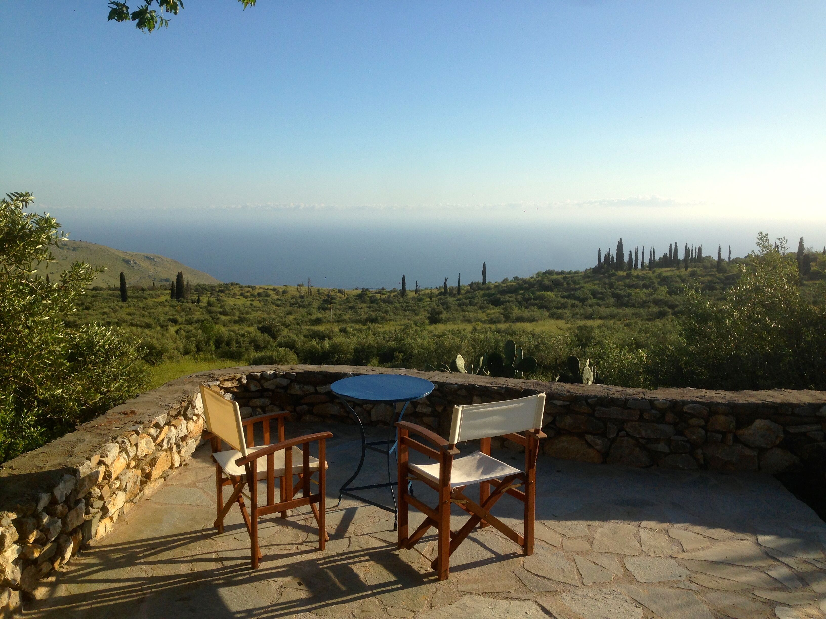 A stone patio with two wooden chairs and a table, overlooking a coastal landscape with greenery and cypress trees.