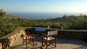 Um pátio de pedra com duas cadeiras de madeira e uma mesa, com vista para uma paisagem costeira com vegetação e ciprestes.