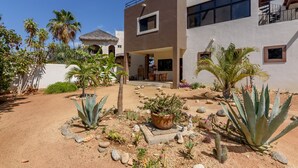 Un edificio moderno de dos pisos con un patio que presenta una variedad de plantas del desierto y una estructura con techo de paja.