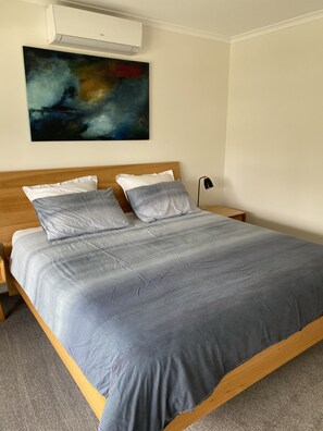 A neatly made bed with a grey duvet and pillows, a wooden headboard, and a large abstract painting on the wall. - Dare St Beach Apartment (Ocean Grove)