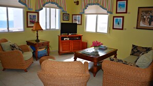 A living room with wicker furniture, a flat-screen TV, and framed pictures on the wall. - Seaside Splendor:  2-3 BR w/ Expansive Deck, Panoramic Views, AC, Wifi, & Pool (Sandy Hill Bay)