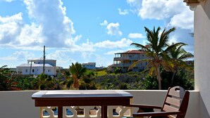 A balcony with a table and chairs, overlooking a coastal area with buildings and palm trees. - Seaside Splendor:  2-3 BR w/ Expansive Deck, Panoramic Views, AC, Wifi, & Pool (Sandy Hill Bay)