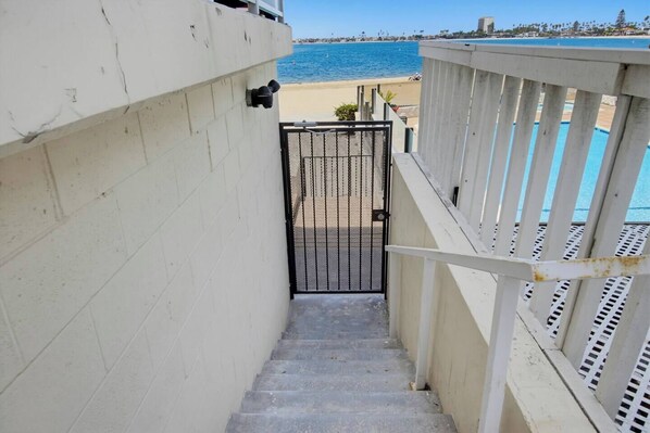 Un escalier en béton mène à une entrée fermée par un portail, d’où l’on a une vue sur la plage et l’océan.