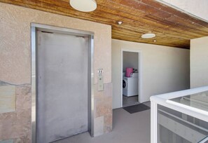 Un couloir avec une porte de monte-charge fermée, une buanderie avec un lave-linge et un escalier menant à l’étage supérieur.