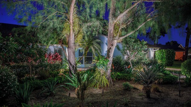 A landscaped garden with various plants and trees, a building with a glass door, and a house in the background. - Oasis in the heart of Equestrian country (Wellington)