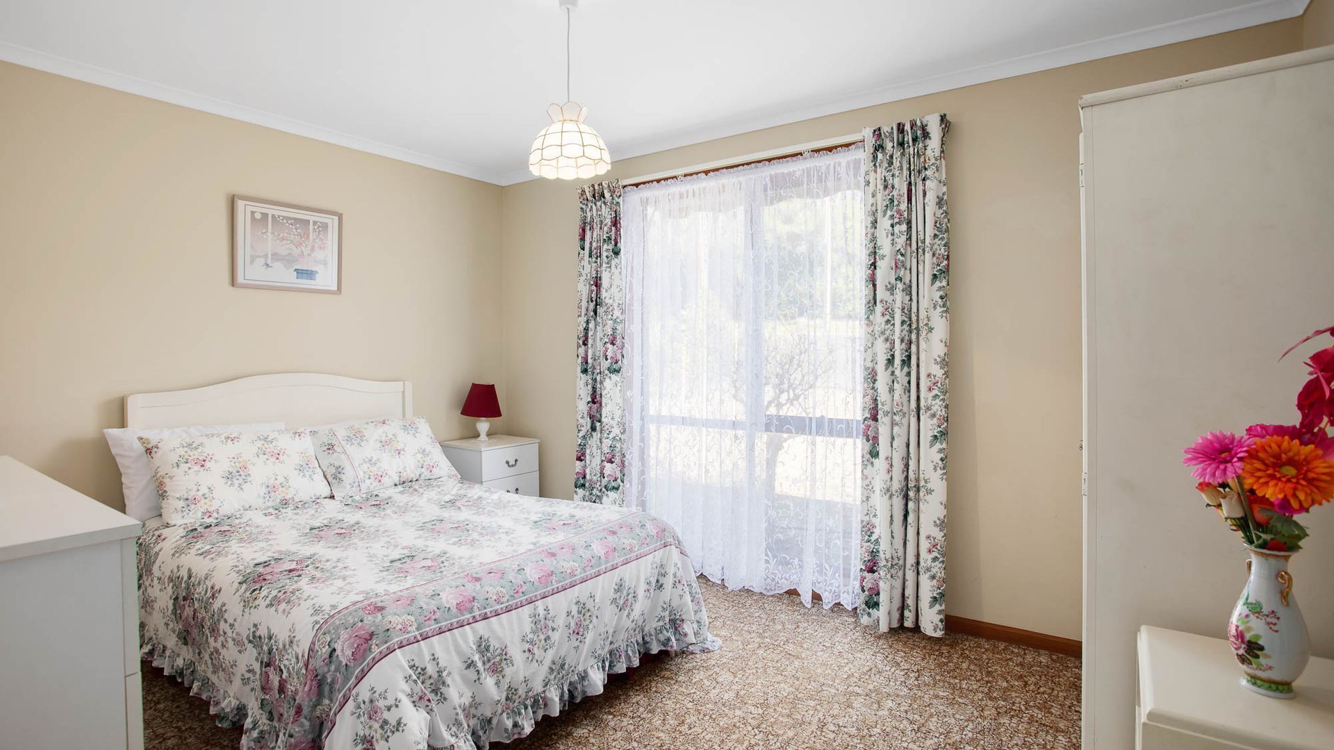 Ein Schlafzimmer mit blumigem Bettbezug, einem Nachttisch mit Lampe, einem Fenster mit floralen Vorhängen und einer Vase mit Blumen auf einem Beistelltisch.