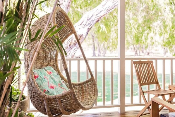 A wicker hanging chair with a cushion, a wooden chair, and a view of the sea through a balcony railing.
